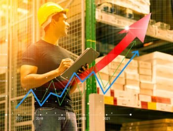 Smart Indian engineer man wearing safety helmet doing stock tick check and cardboard stock product management in factory warehouse background and profit chart grow up